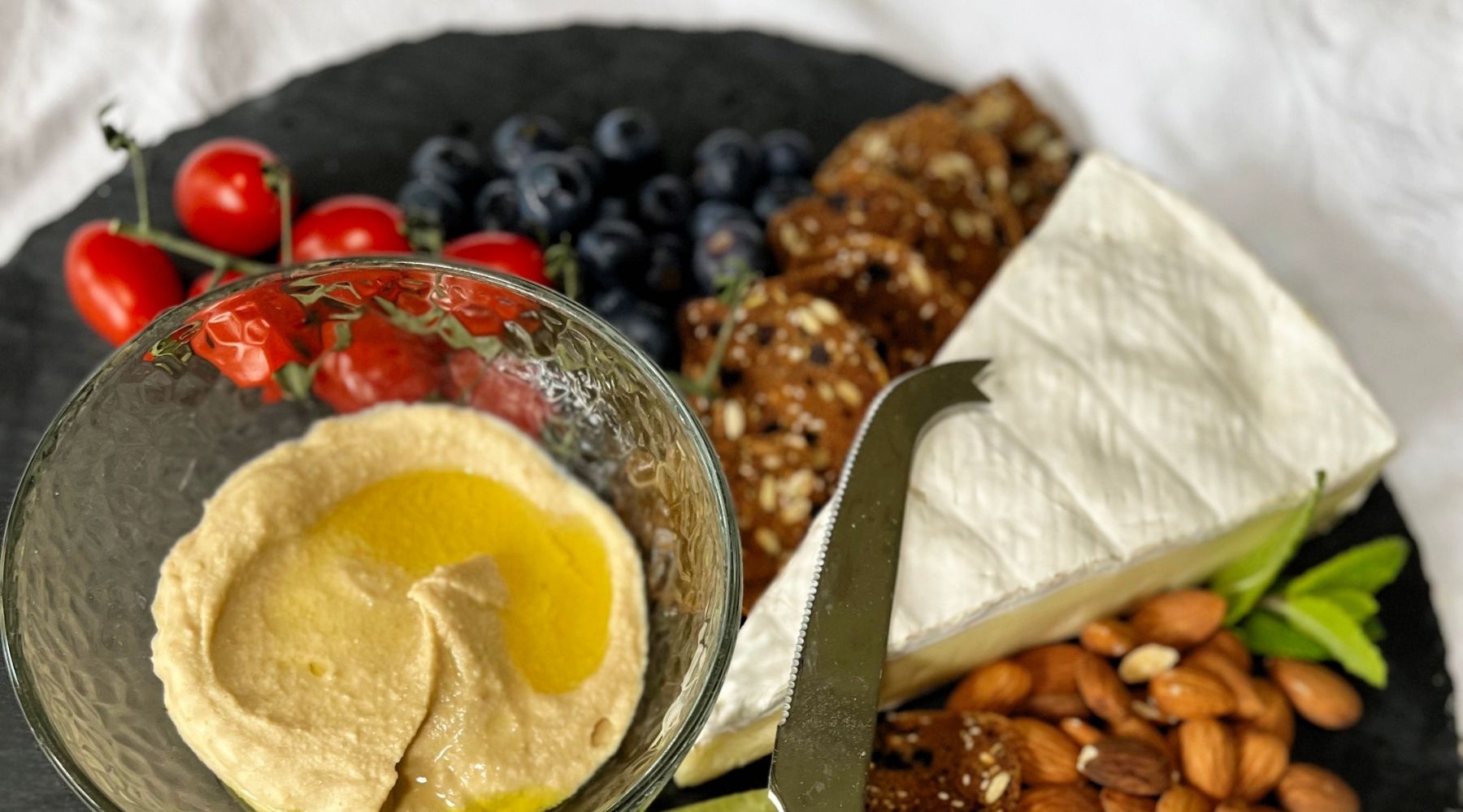 A gluten-free cheese board made up of a round charcoal slate, wild blueberry walnut crisps, brie, almonds, grapes, vine tomatoes and hummus. 