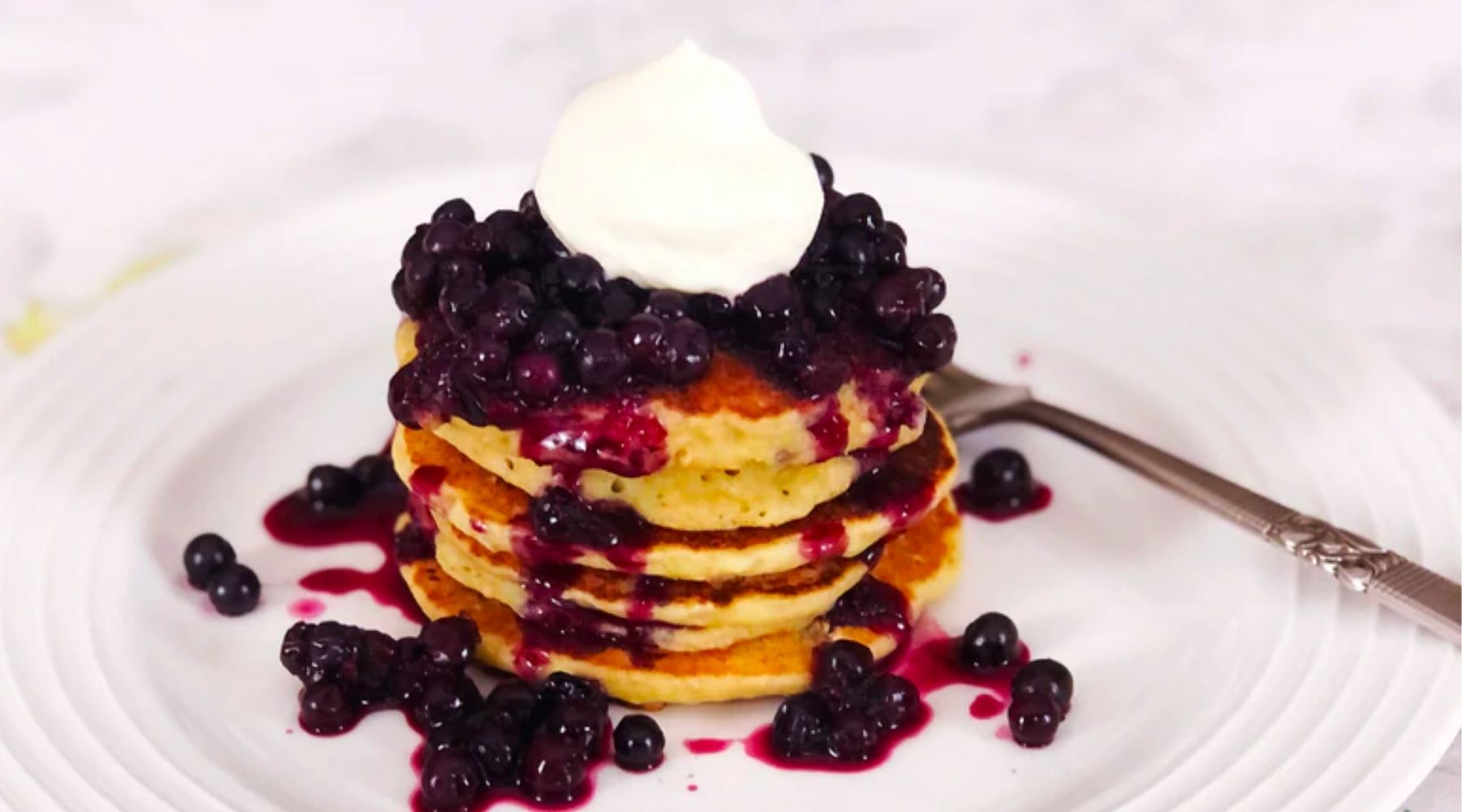 Wild Blueberry Lemon Ricotta Pancakes on a white plate with whipped cream and a fruit patterned table cloth.